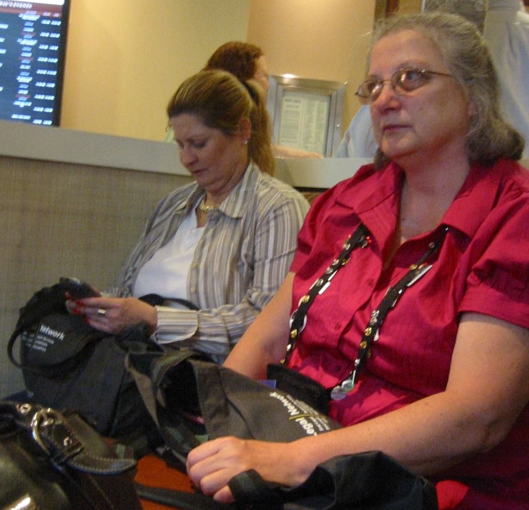 Two women sitting at a table in an airport, awaiting their flight. One is using a cell phone, and the other is looking forward, wearing glasses and a red shirt.