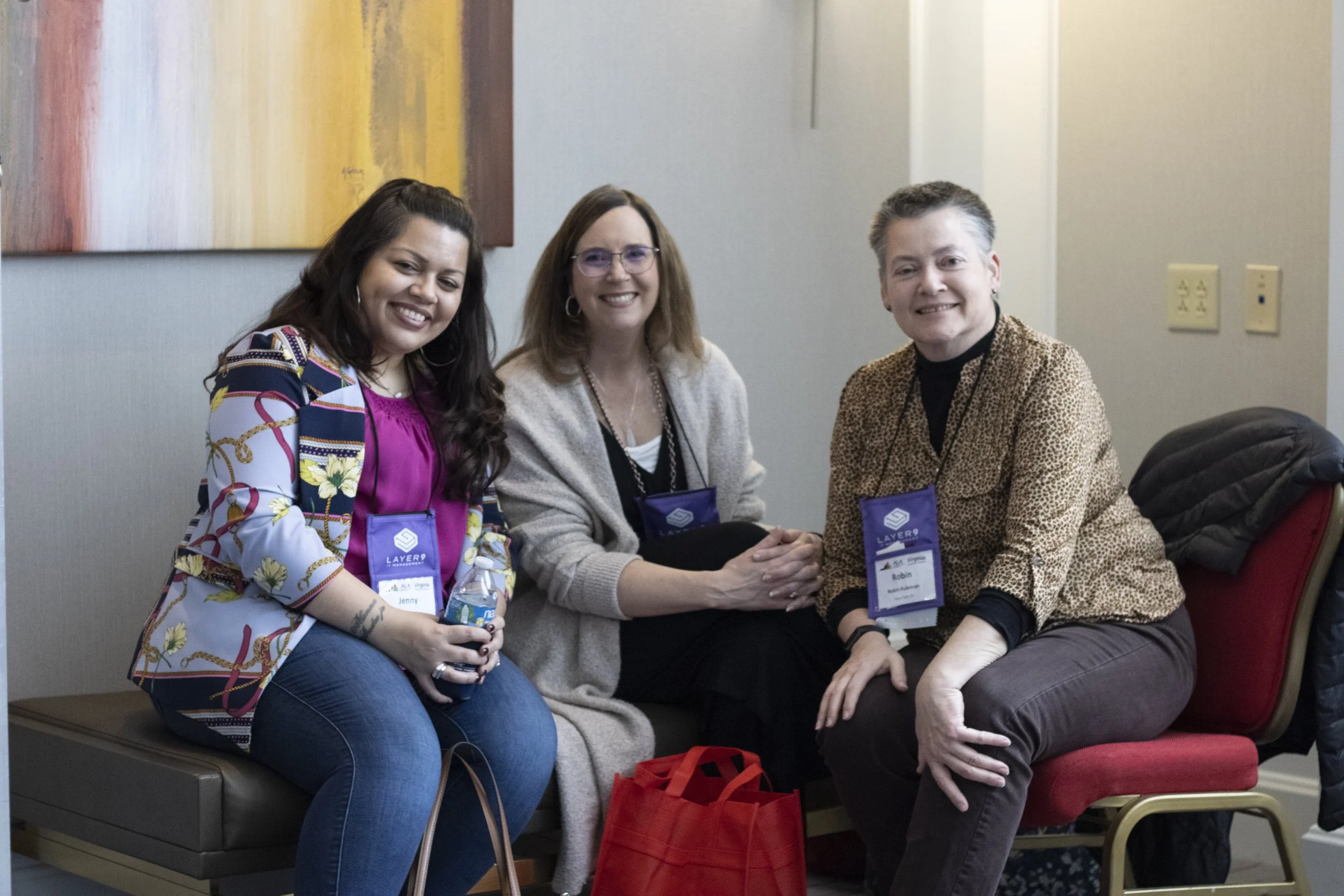 Three women sitting in a row, smiling at the camera. All are wearing conference badges and are seated on a bench with a gray wall and colorful artwork behind them.