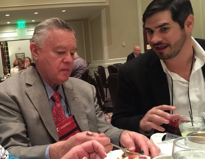 Two men sitting at a table engaged in conversation, one older with gray hair and a checkered blazer, the other younger with dark hair and a black blazer, at a formal event in a banquet hall.