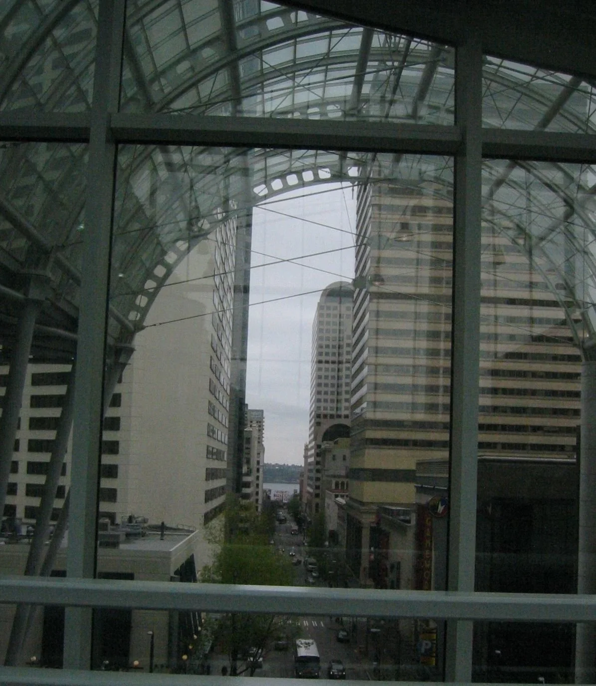 City view through glass walls featuring tall buildings on either side and a street below with vehicles, with a body of water in the distance.