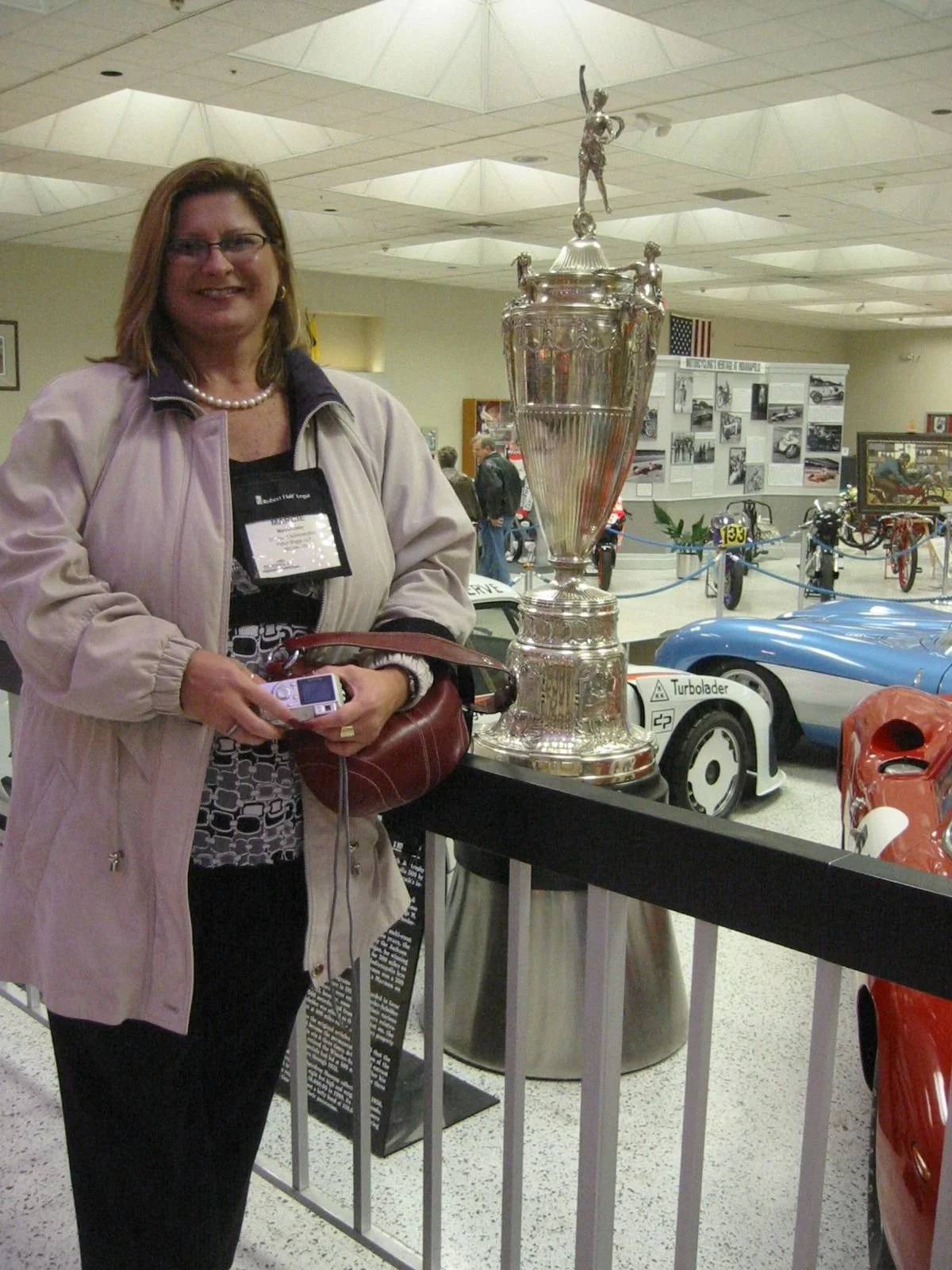 A woman with glasses and a pearl necklace standing next to a large silver trophy in a museum filled with vintage cars and photos.