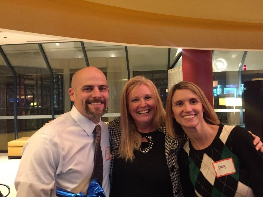 Three people smiling and standing together indoors at night, with a glass wall and city lights in the background. One man and two women, one of the women has a name tag that reads 'Stacy'.