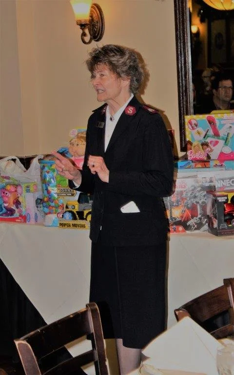 Woman in a black uniform with a red S on her collar standing in a room with toys on a table behind her.