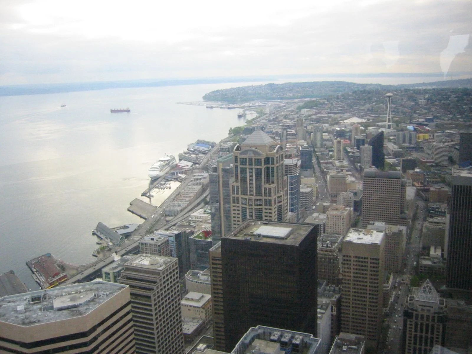 A view of downtown Seattle from a high-rise, showcasing tall buildings, a waterfront with ships, and the Space Needle in the distance under overcast skies.