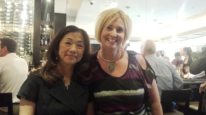 Two women smiling at a restaurant or bar, with other people in the background.