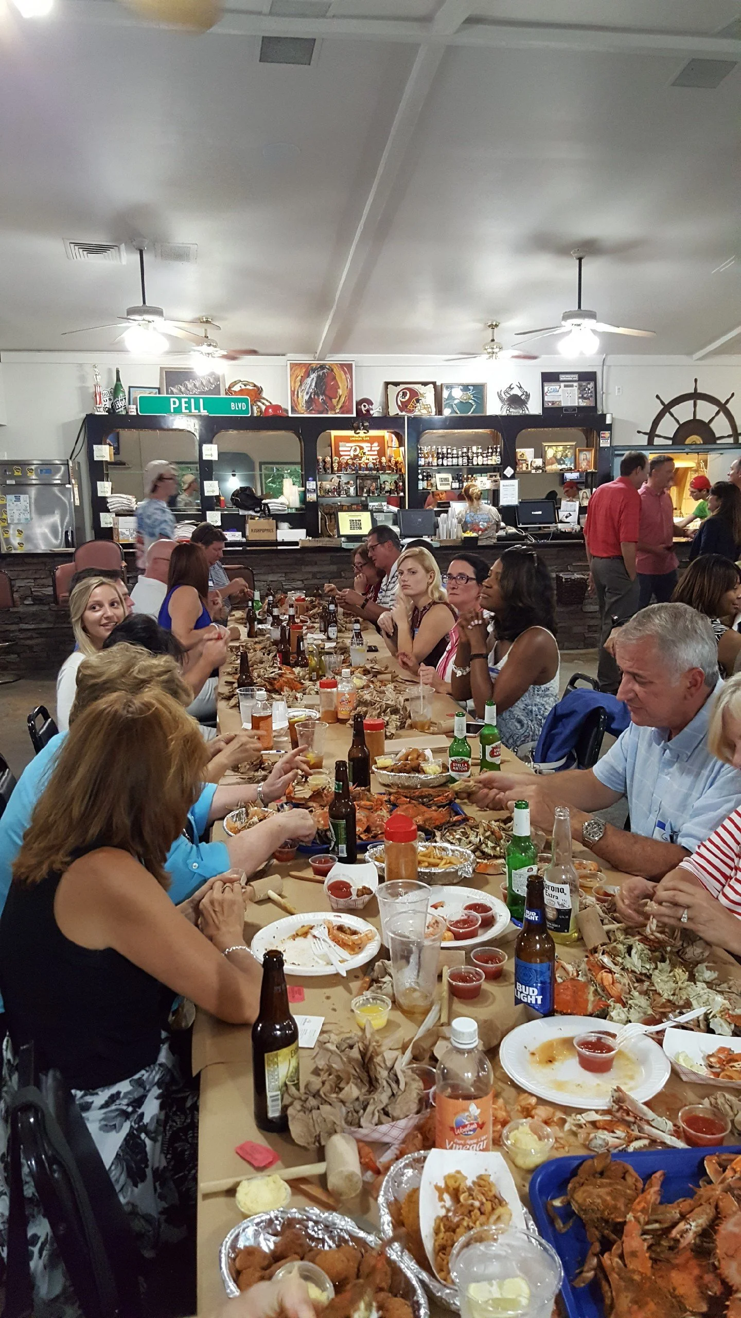 A long table filled with people dining, eating seafood and drinking beer at a crowded restaurant. Food and drinks are scattered across the table, with some plates of seafood, including lobster and fried items, visible.