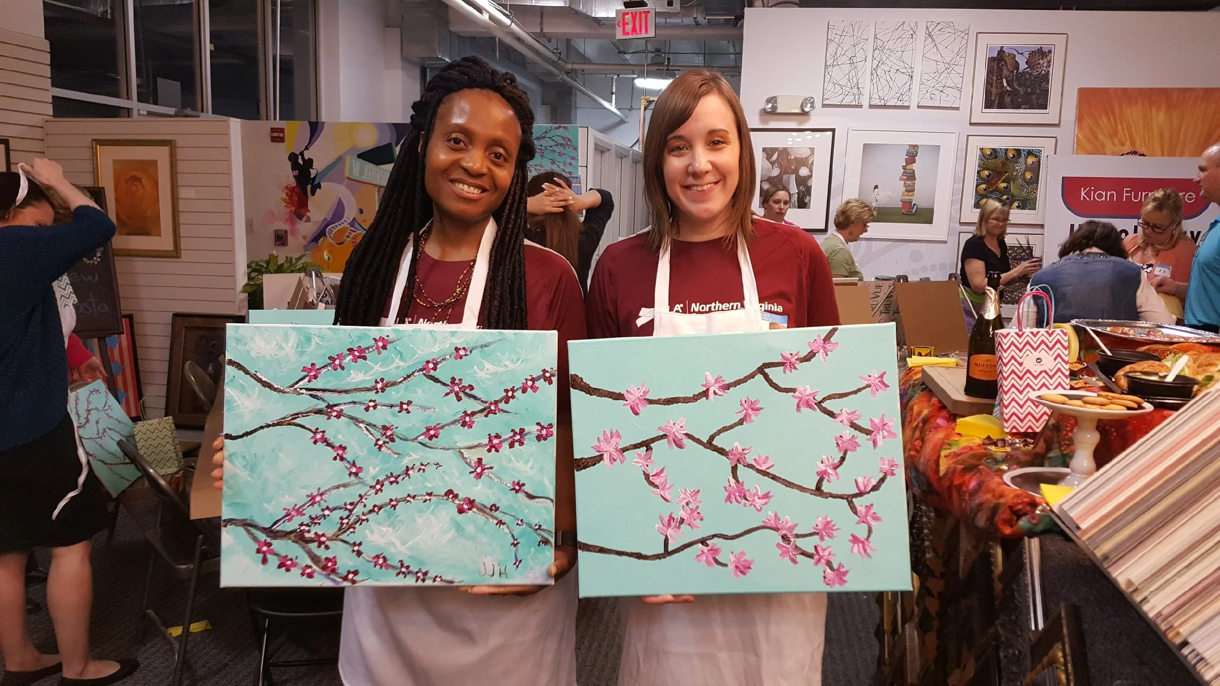 Two women standing in an art gallery or craft fair, each holding a painting of pink cherry blossoms on a blue background. The women are smiling and wearing maroon shirts and aprons.