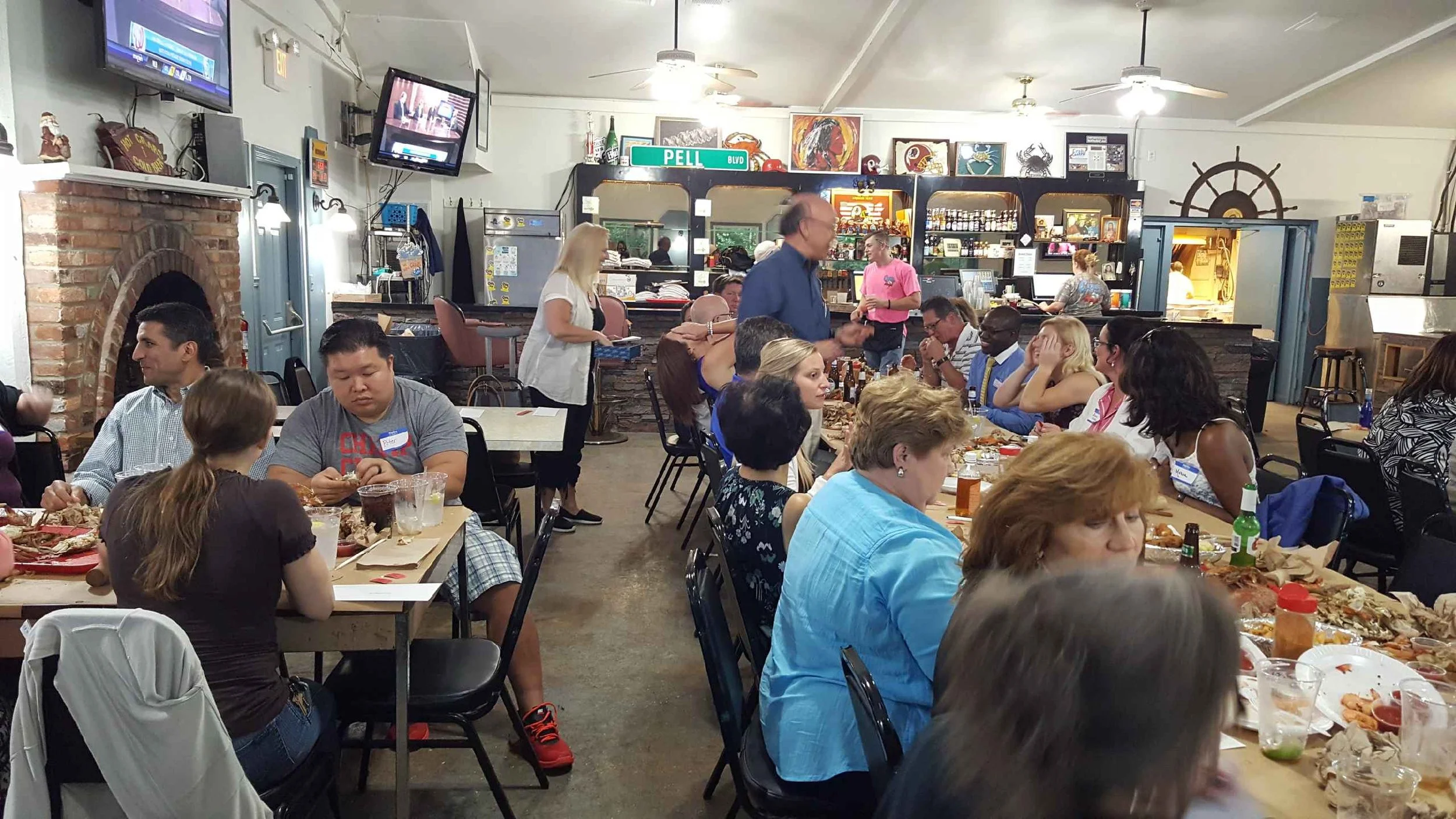People seated at long tables enjoying a meal in a cozy restaurant with decorative artwork, a brick fireplace, and a bar area in the background.
