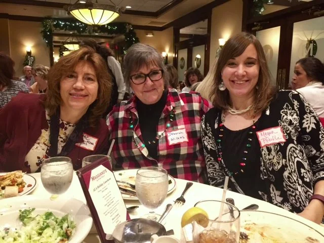Three women sitting at a dinner table, smiling, at an indoor event or banquet.
