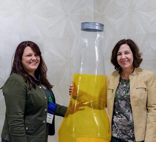 Two women smiling and holding a giant plastic bottle filled with a yellow liquid, standing in front of a patterned wall.
