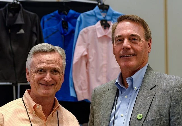 Two men smiling and posing for a photo in front of a display of men's dress shirts in various colors, including blue and pink.