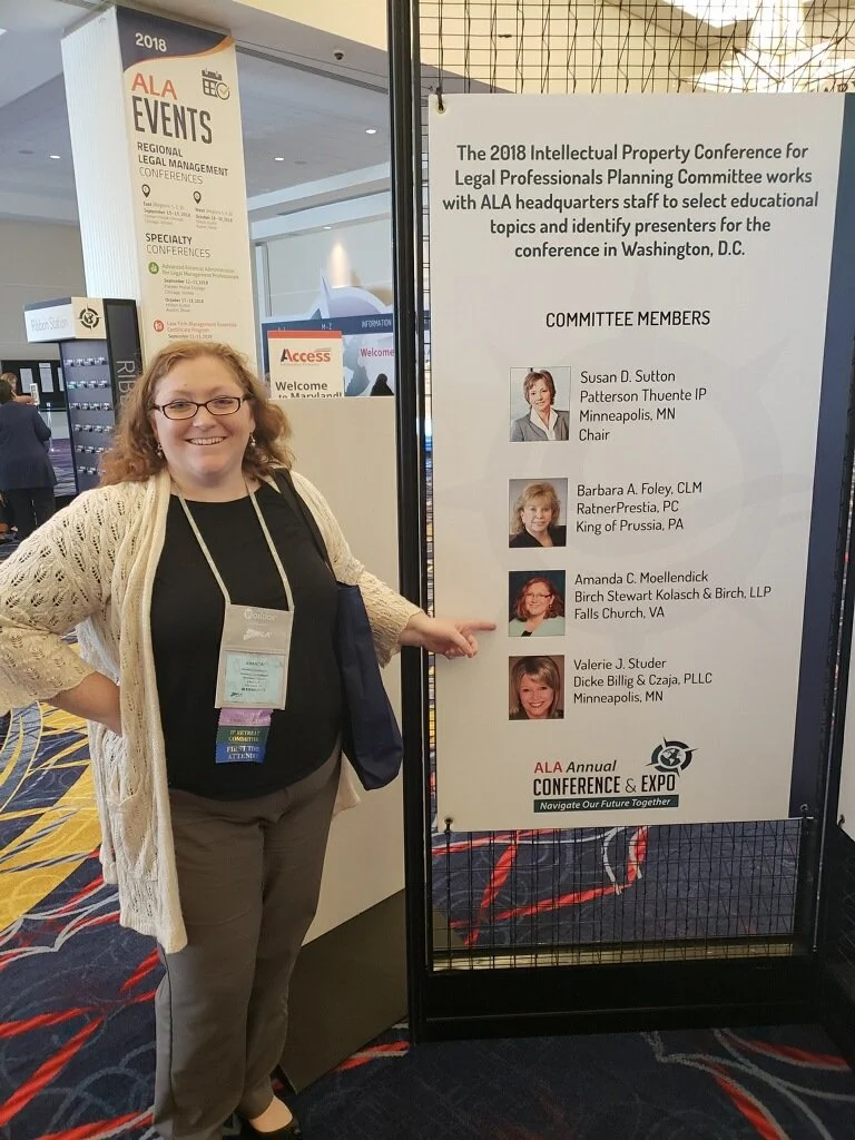 Woman standing next to a conference sign at the 2018 Intellectual Property Conference for Legal Professionals. The sign displays committee members' names and photos, including Susan D. Sutton, Barbara A. Foley, Amanda C. Moellendick, and Valerie J. S