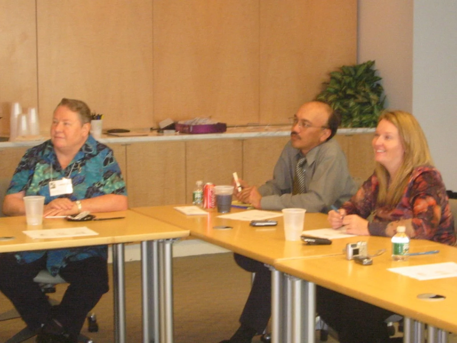 Three people sitting at a conference table in a meeting room, with papers, beverages, and electronic devices on the table.