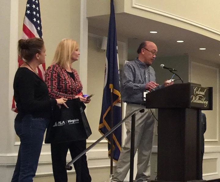 Three women and one man standing at a podium during an indoor event, with American and Virginia flags in the background; the man is speaking into a microphone, while the women are listening.