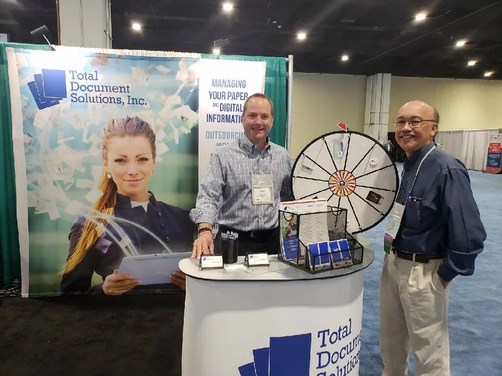 Two men standing at a booth with a large backdrop featuring a woman holding a tablet and the logo of Total Document Solutions, Inc. The booth has a spinning wheel, informational materials, and a table with business cards.