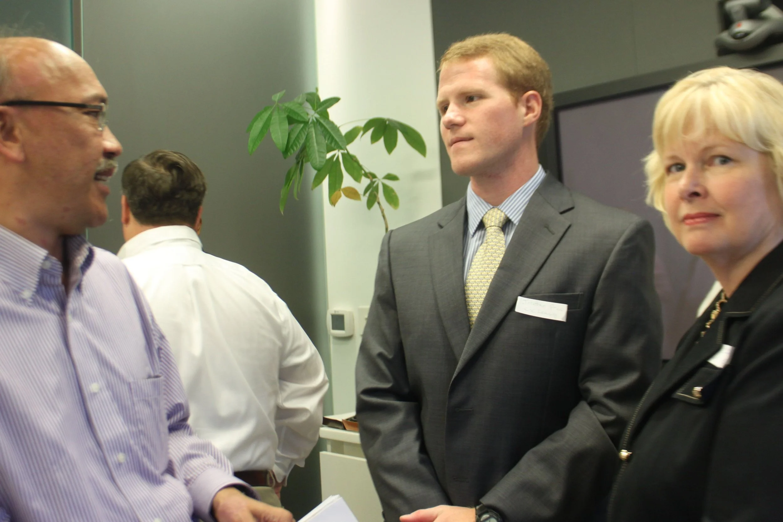 People engaging in conversation in a professional setting, including a man in a suit and a woman with blonde hair, with a green plant in the background.