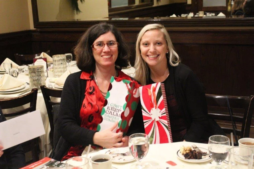Two women sitting at a Christmas dinner table, smiling, holding holiday-themed gift bags, with plates of food and drinks in front of them.
