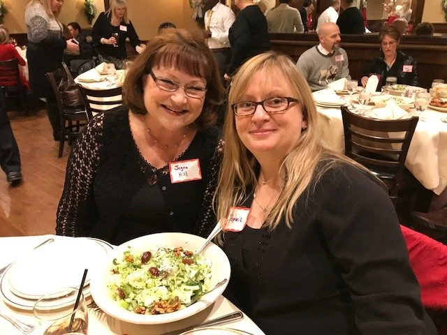 Two women sitting at a banquet table smiling, with a large salad in front of them. The woman on the left has short brown hair, glasses, and is wearing a black top with a name tag that says "Joyce Hull." The woman on the right has long blonde hair, gl