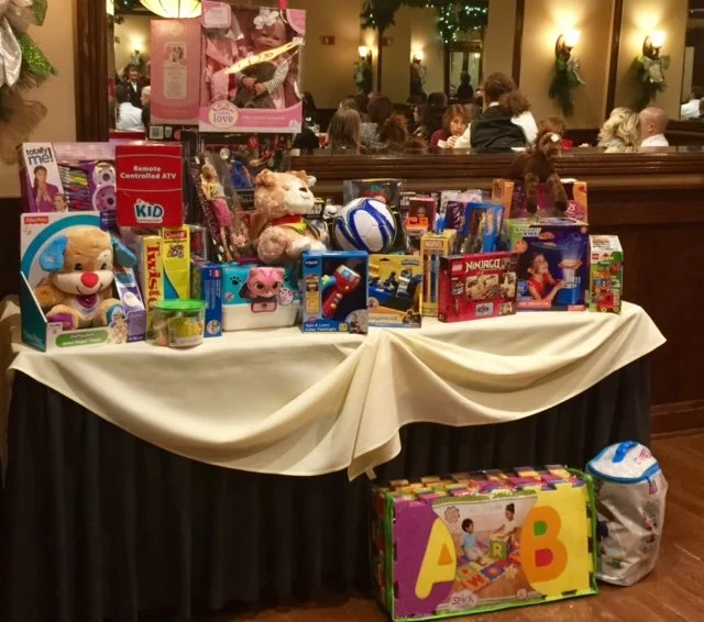 Table filled with various toys, including plush animals, a soccer ball, and children's games, set in a restaurant with people dining in the background.