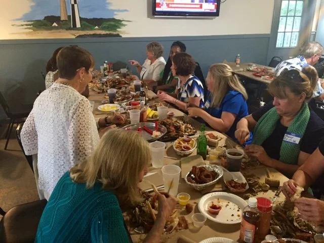 Group of women enjoying a meal together at a long table, sharing food and drinks in a casual dining setting.