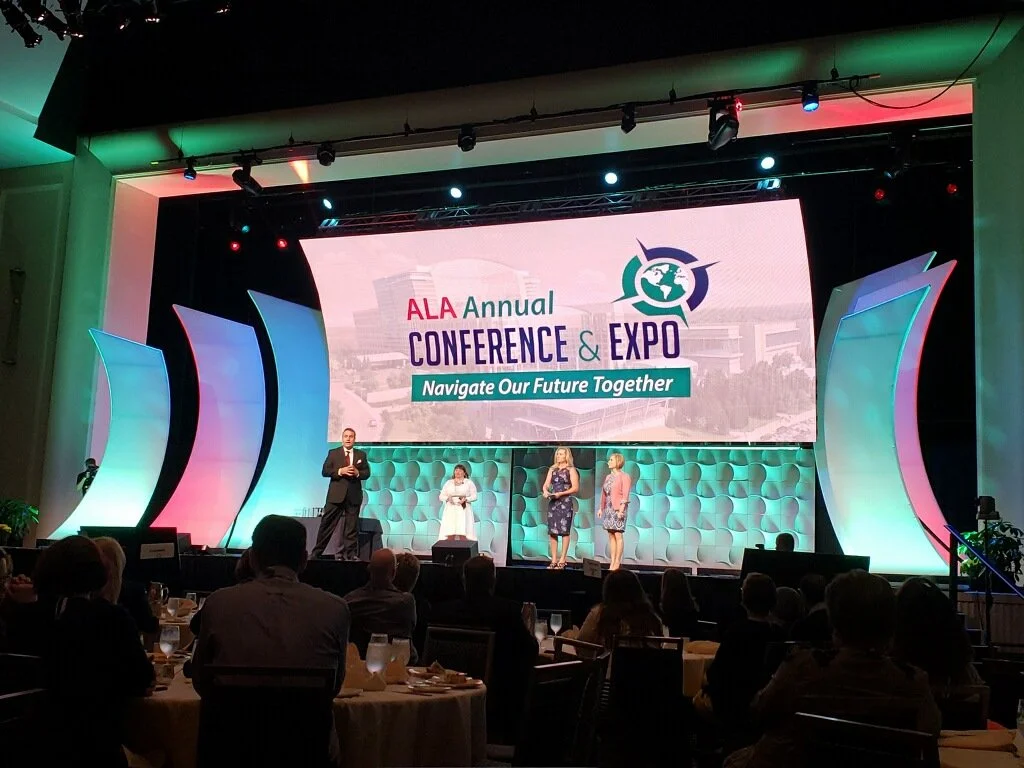 Stage at the ALA Annual Conference and Expo with a large screen displaying the event logo and slogan, surrounded by colorful lighting and decorated panels, with attendees seated at round tables.