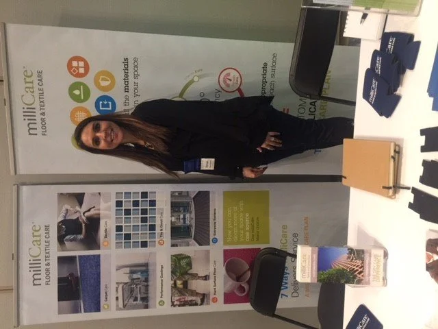 A woman standing next to a table with brochures and items, in front of a display stand for Milliken Floor & Textile Care at a trade show or conference.