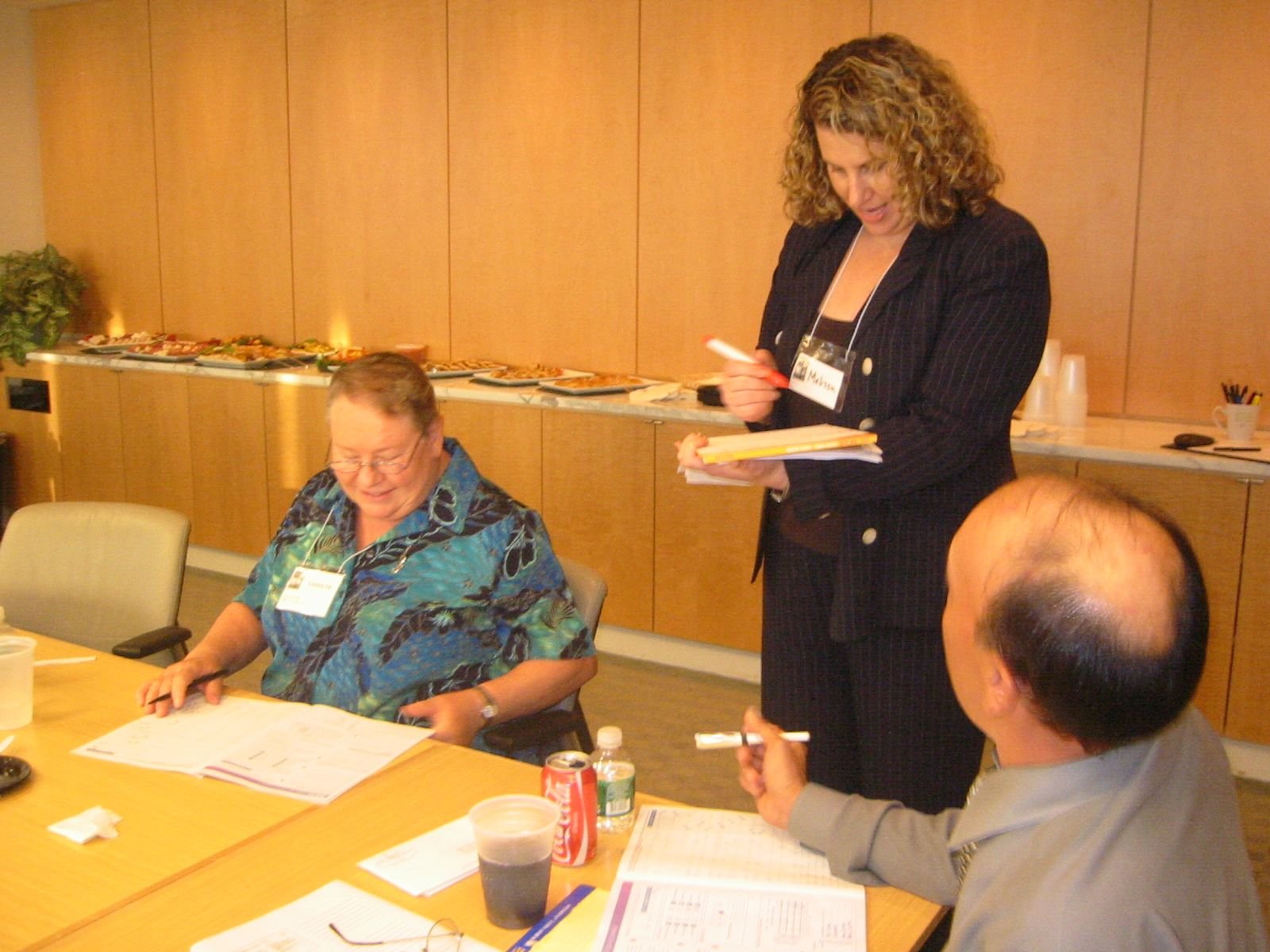 Group of three people in a meeting room with a buffet of food in the background. Two men and a woman are engaged in discussion.