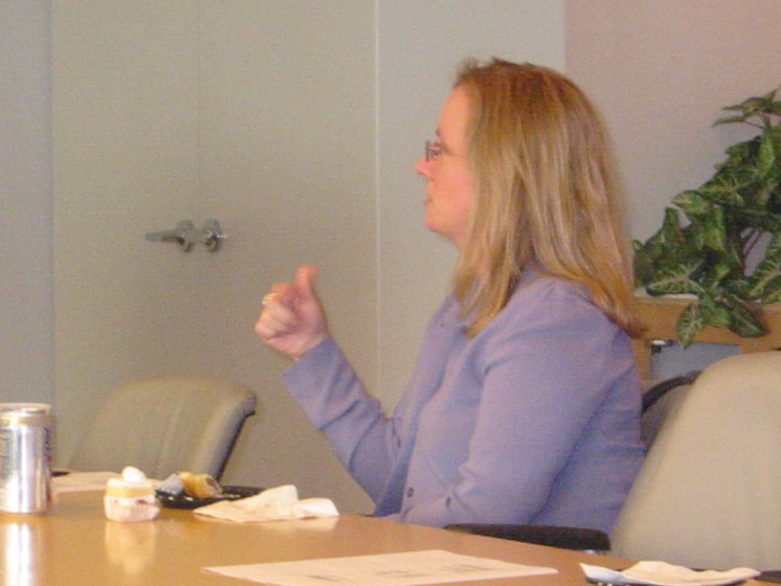 A woman with reddish hair wearing glasses and a purple blazer, sitting at a conference table in a meeting room, gesturing with her right hand, with a plant and beige chairs in the background.