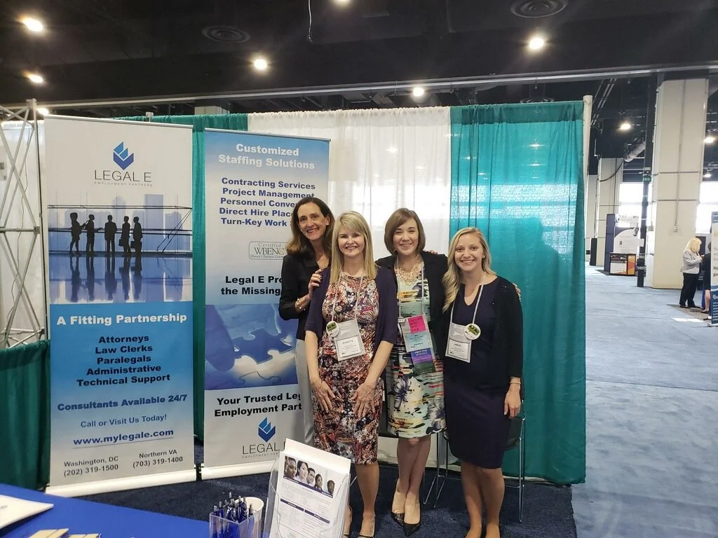 Four women standing in front of booth banners at a conference, smiling for the photo.