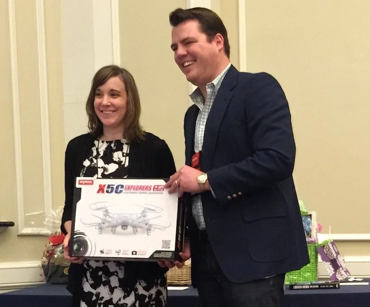 A woman and man smiling, holding a drone box, at an indoor event with gifts and decorations in the background.