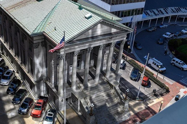A historic building with large columns and stairs, an American flag flying, and a parking lot surrounding it.