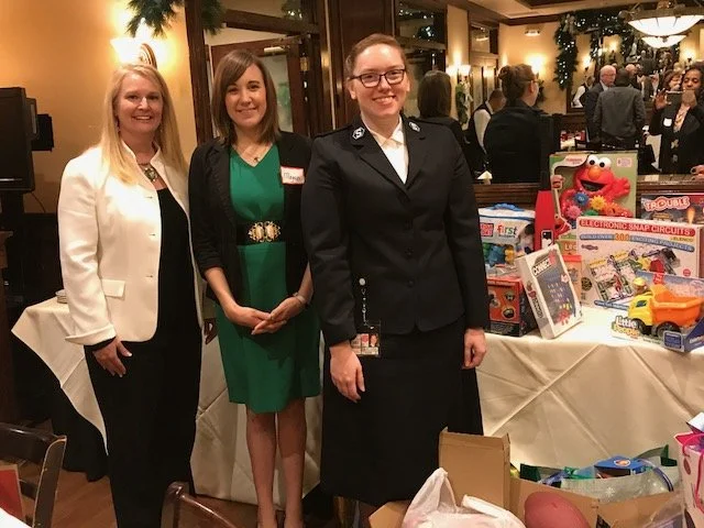 Three women standing next to a table with toy prizes in a decorated indoor setting, possibly during an event or fundraiser.