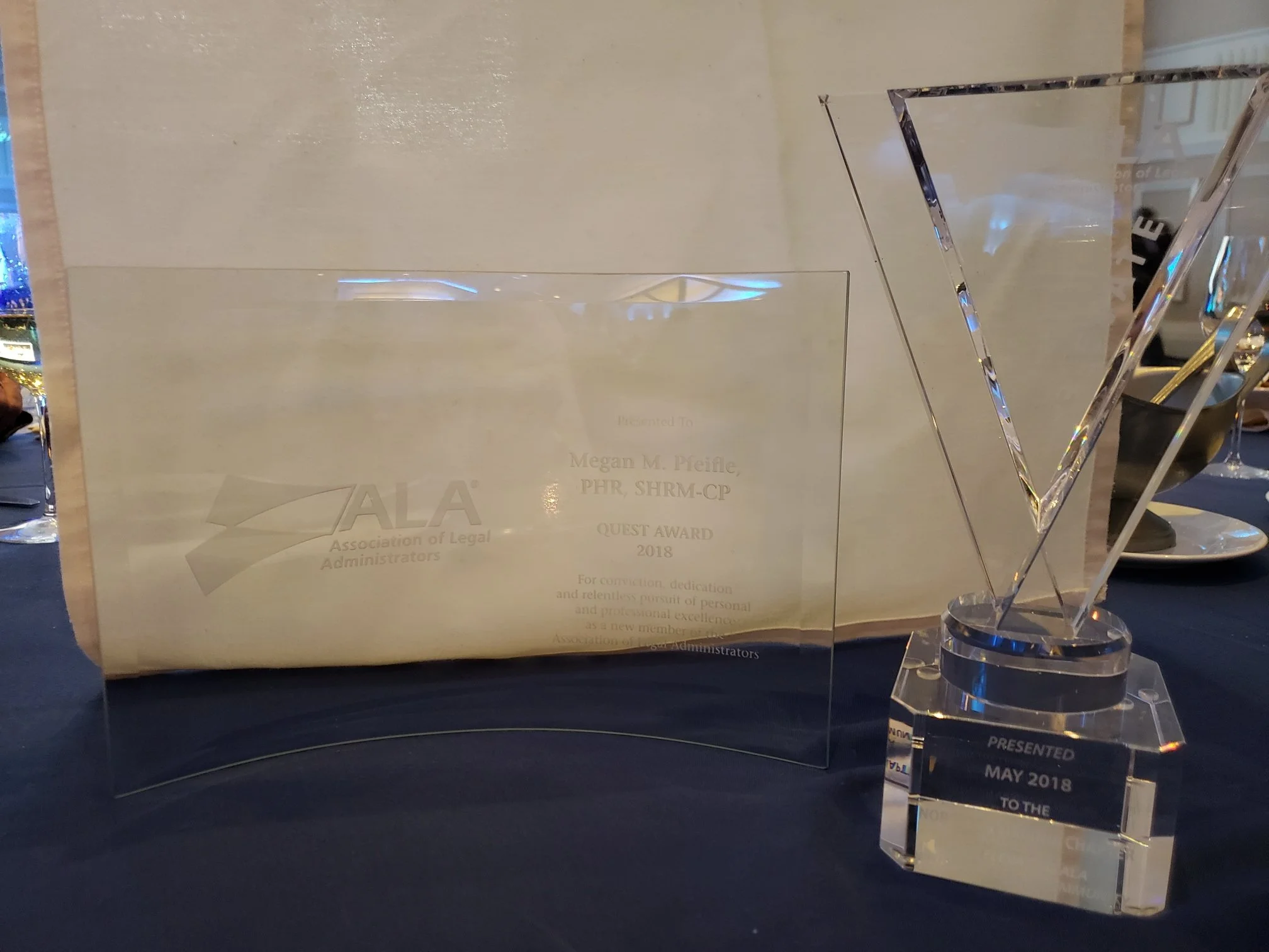 A clear acrylic award plaque and a crystal award on a black surface with a beige wall in the background. The plaque has text including 'Presented To Megan M. Pfeifle, PHR, SHRM-CP, Quest Award 2018.'