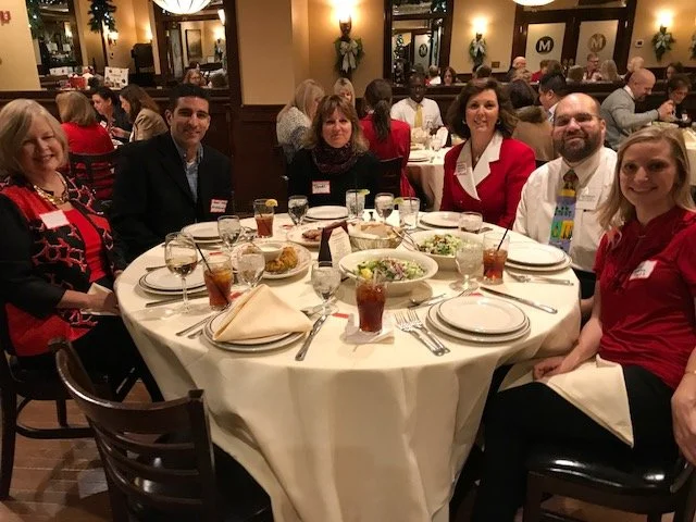 Six people sitting around a round dining table with plates, glasses, and food in a restaurant.