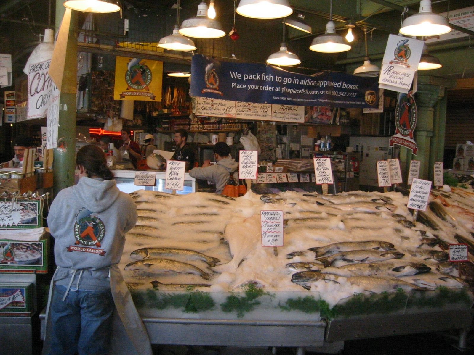 Fish market with piles of fish on ice, fishmongers and customers in the background, various signs with prices, and posters promoting shipping and brand.