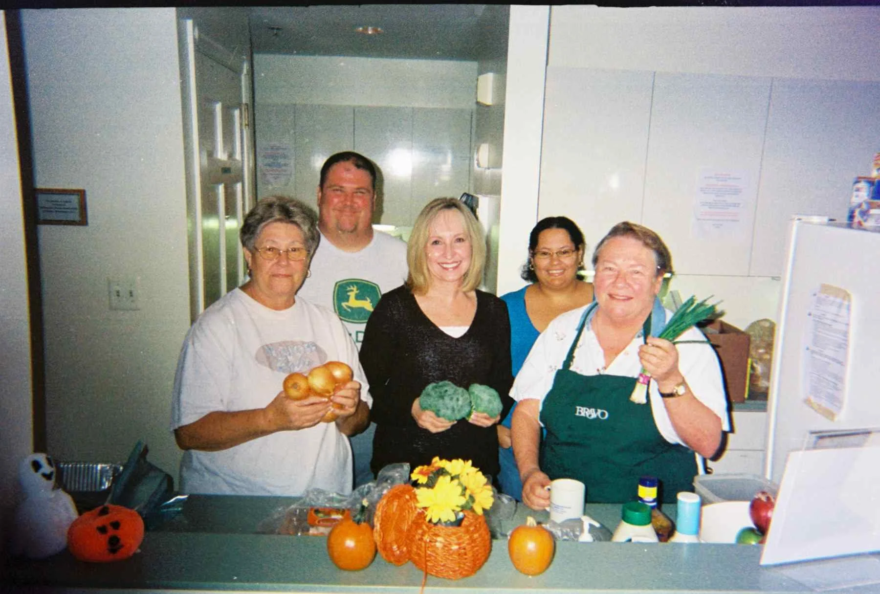 Four women are in a kitchen, holding vegetables and apples, with a decorative pumpkin on the counter, and smiling at the camera.
