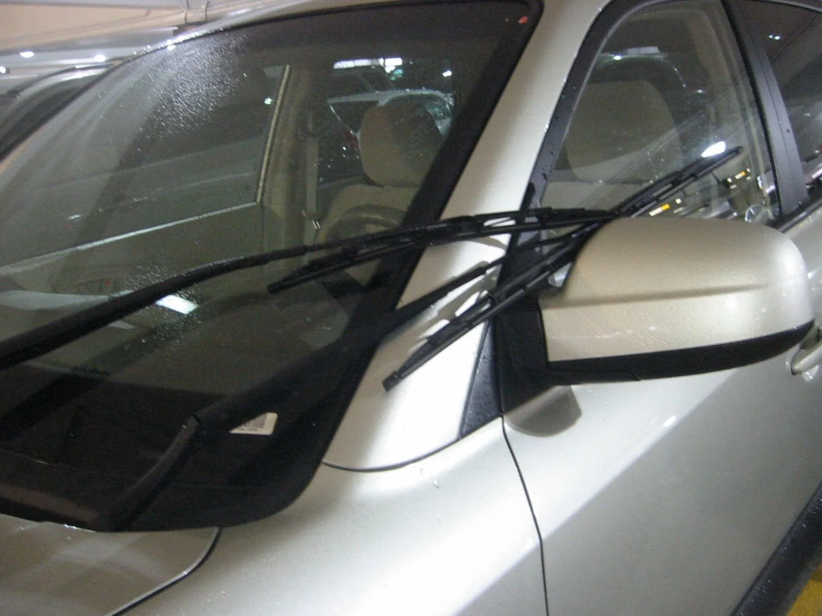 Close-up of a silver/white car window with rain droplets, a side mirror, and windshield wipers in a parking garage.