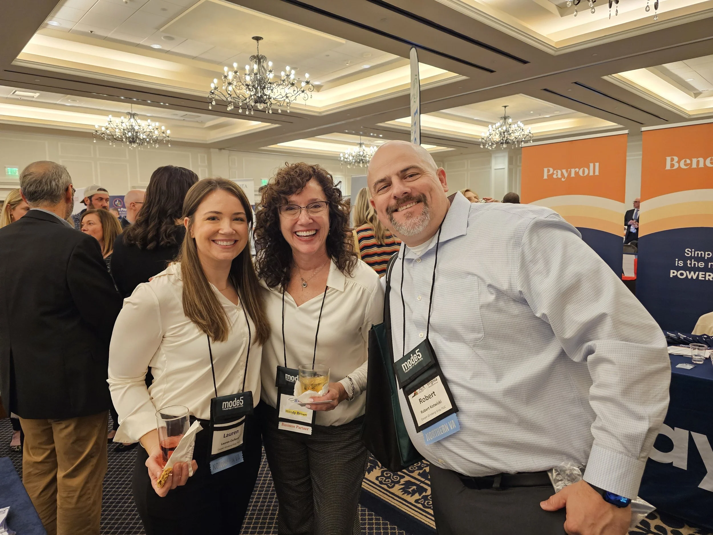 A group of three smiling people, two women and a man, at a professional networking event or conference. They are wearing name badges and are standing in front of a booth with orange banners labeled 'Payroll' and 'Benefits.' The background shows other