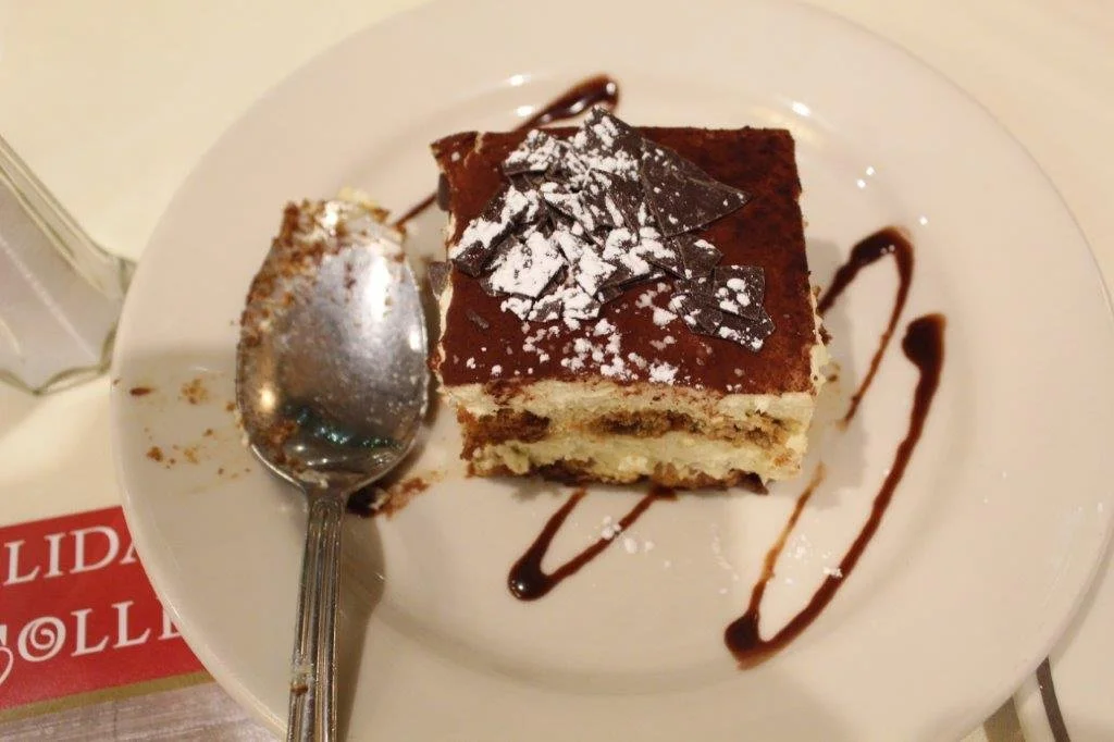 A plate with a slice of tiramisu cake, chocolate shavings, and a drizzle of chocolate sauce, with a spoon resting on the plate.