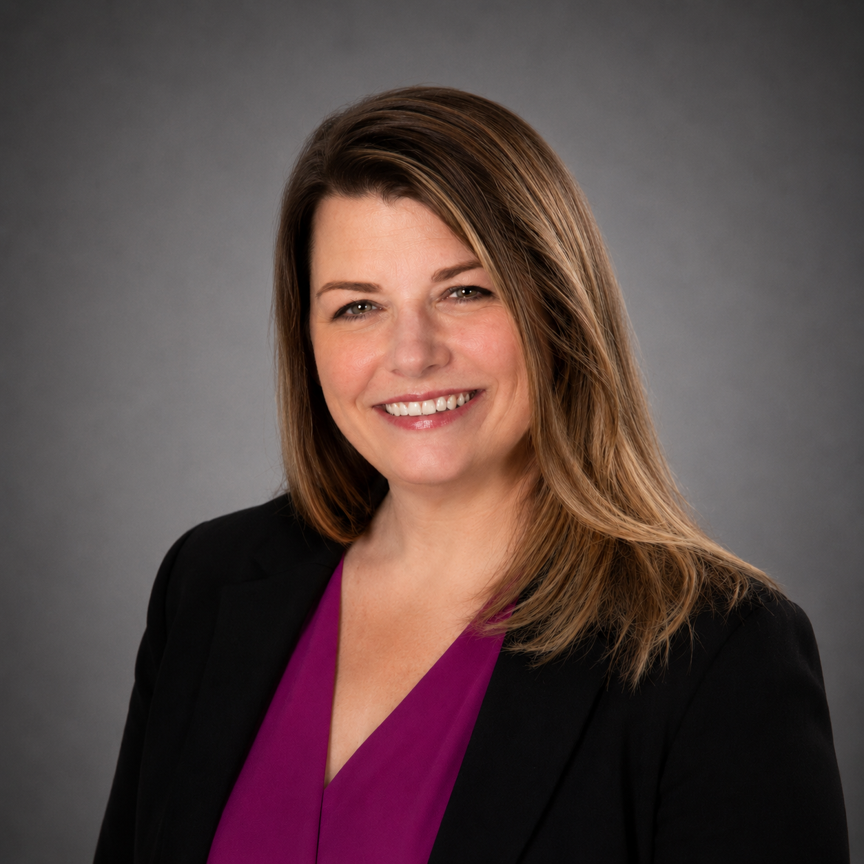A professional woman with shoulder-length brown hair, smiling, wearing a black blazer and a purple top, against a gray background.