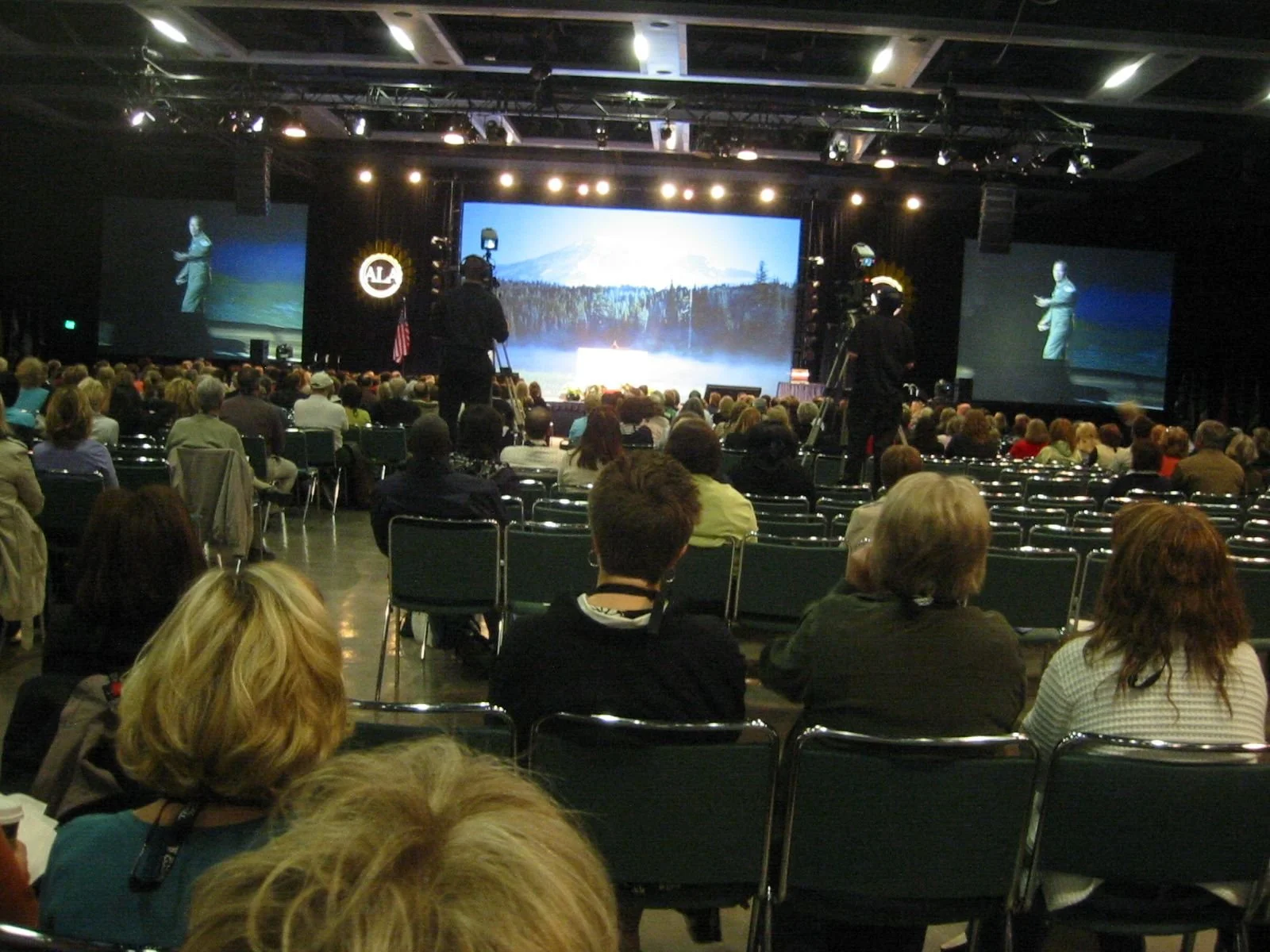 A large audience seated in a conference hall watching a presentation on a stage with multiple large screens and a mountain landscape background.