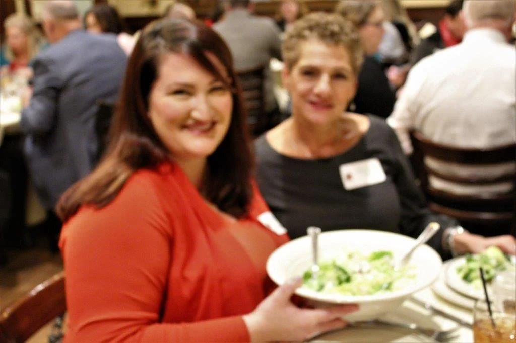 Two women smiling at a dinner table with salad bowls in front of them, surrounded by other diners in a busy restaurant.
