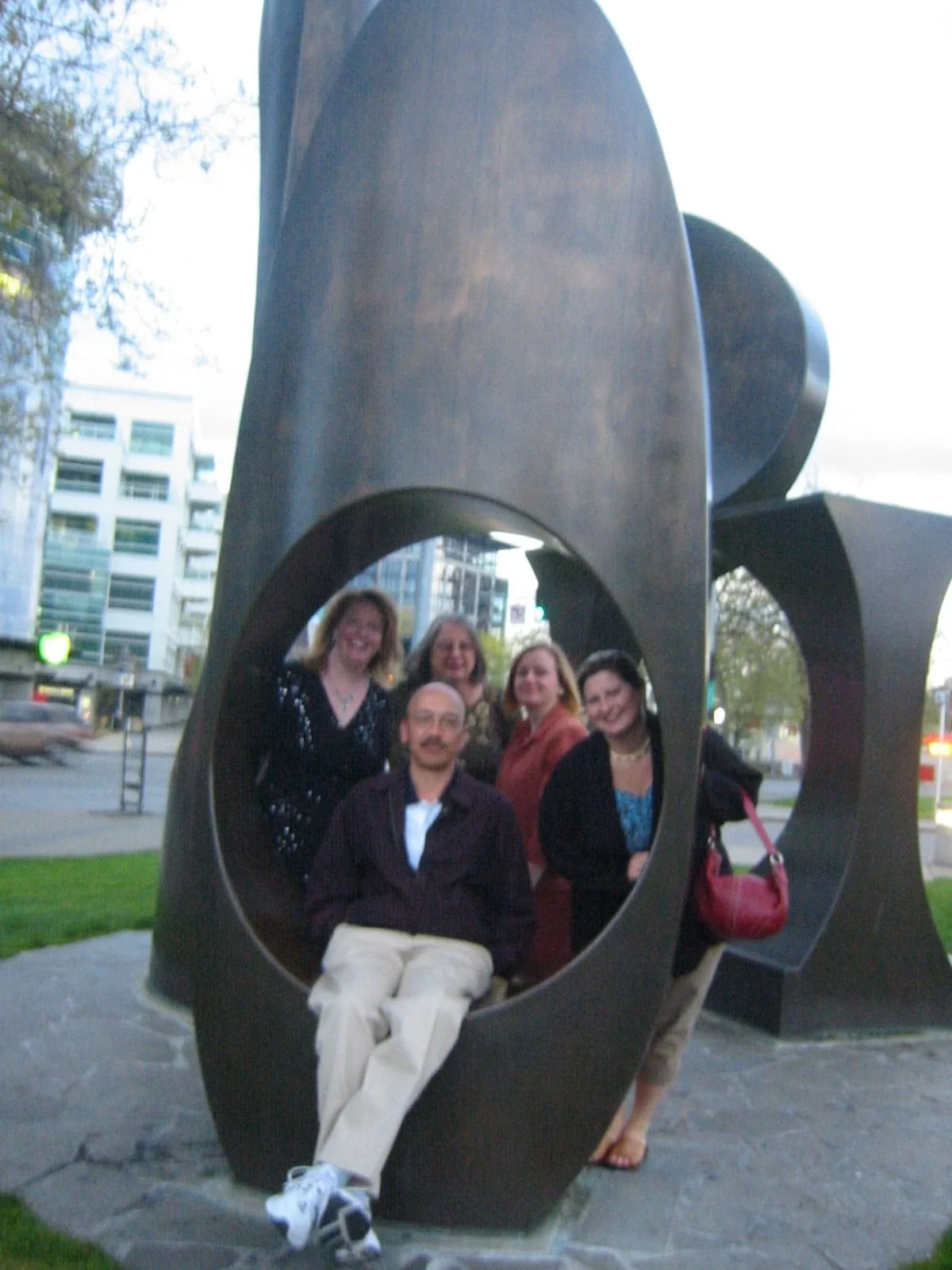 Group of six people posing inside a large abstract metal sculpture with circular cutouts, outdoors in an urban area with buildings, trees, and street in background.
