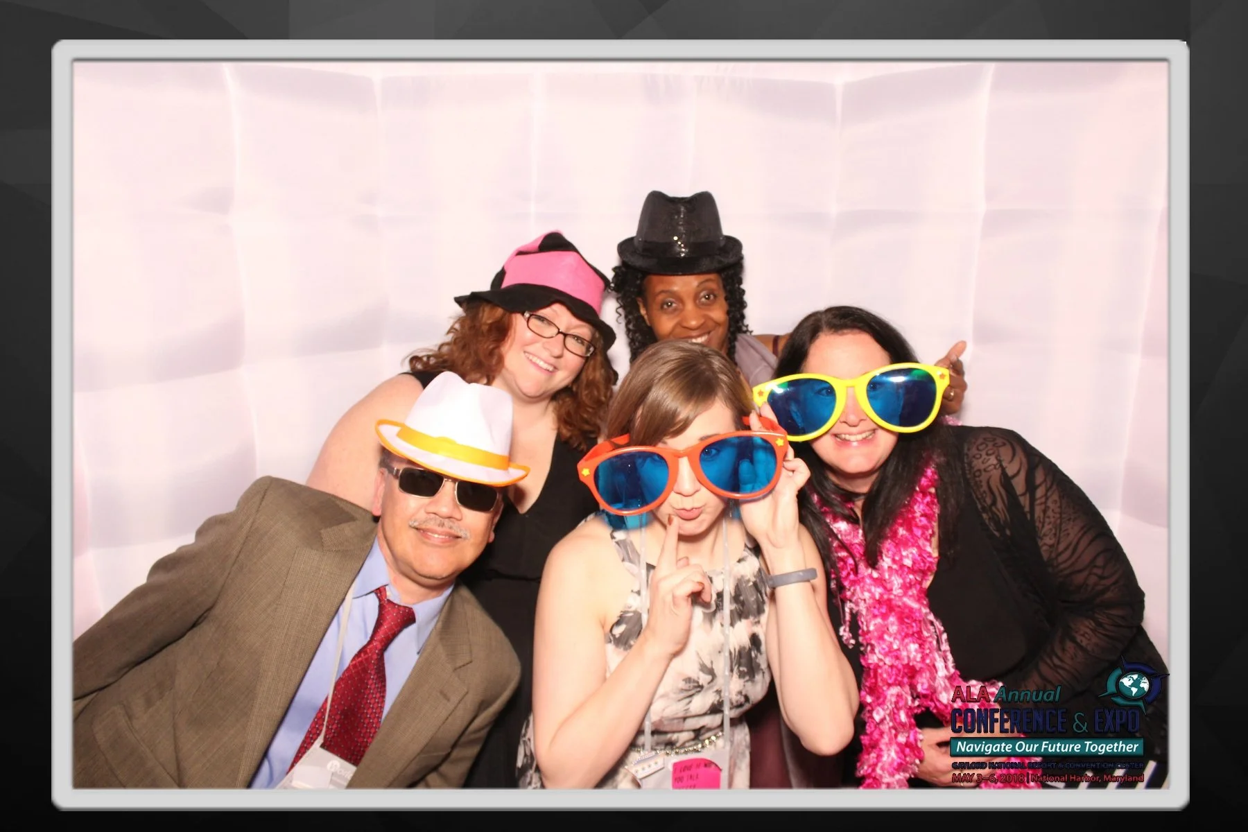 A group of six diverse people, wearing colorful oversized sunglasses and fun hats, smiling and posing for a photo at a conference with a white backdrop. The conference logo and tagline are visible in the bottom right corner.