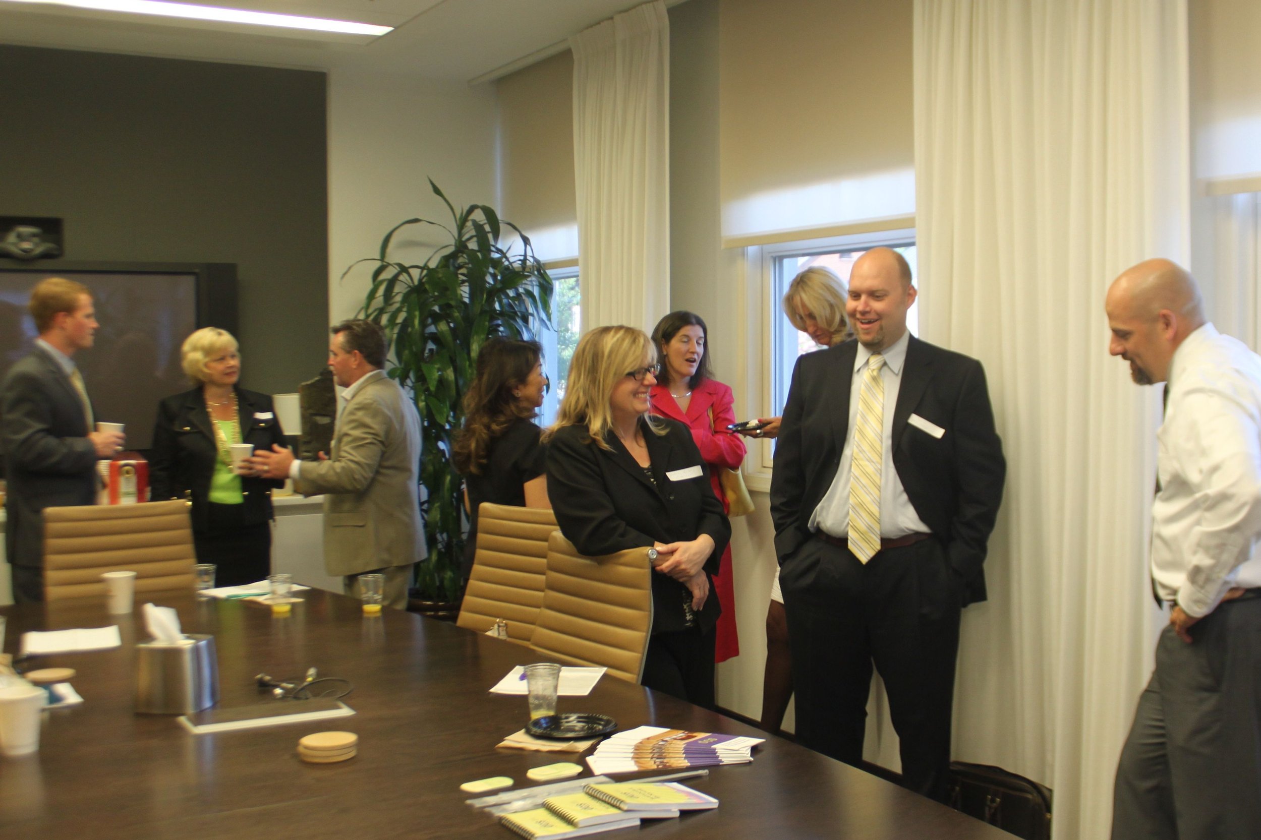 Group of business professionals socializing in a conference room with a large wooden table, notebooks, and refreshments.