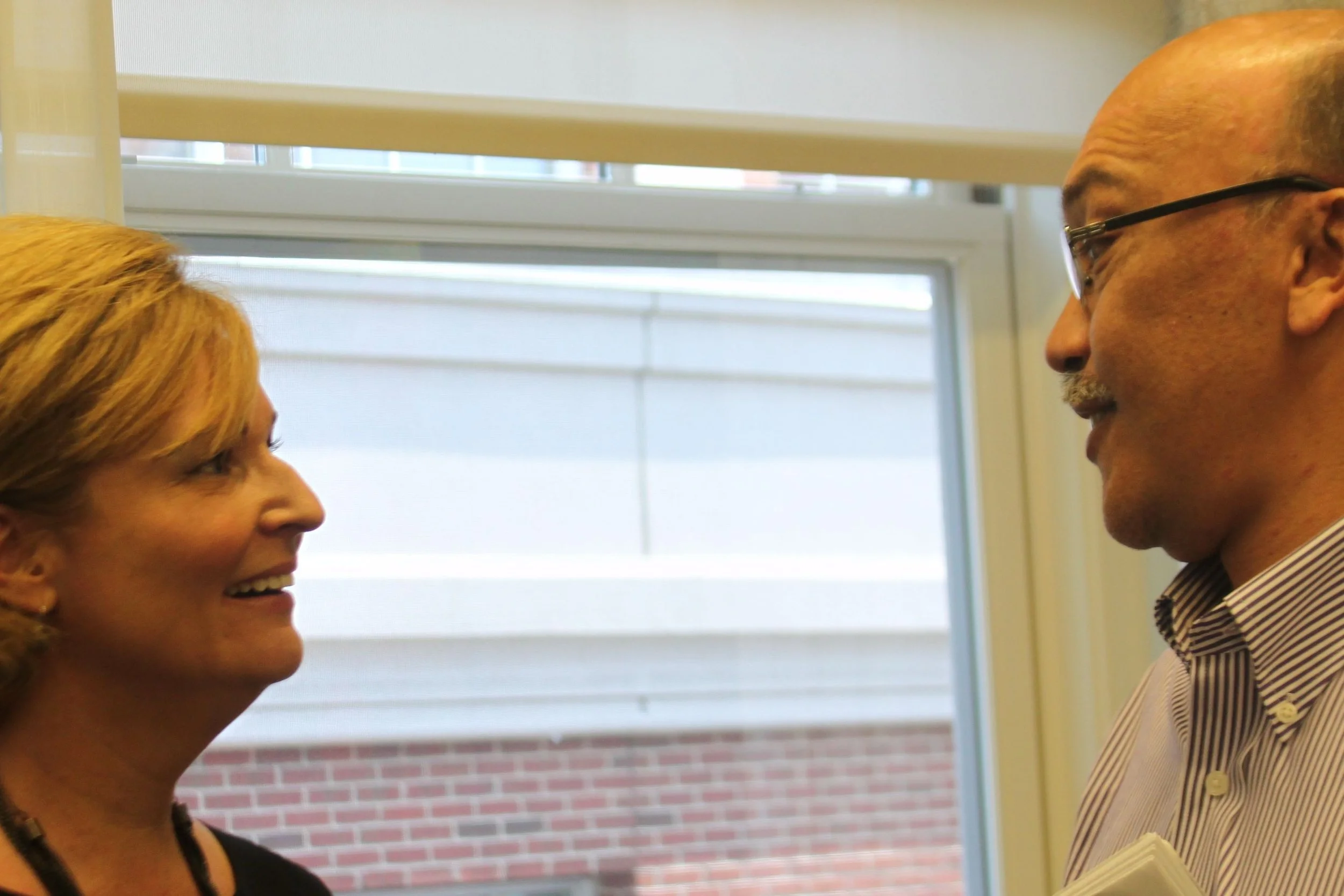 Two people, a woman and a man, are facing each other indoors near a window. The woman has shoulder-length blonde hair and is smiling; the man has a balding head, wears glasses, and is dressed in a striped shirt and blazer. They are engaged in a conve