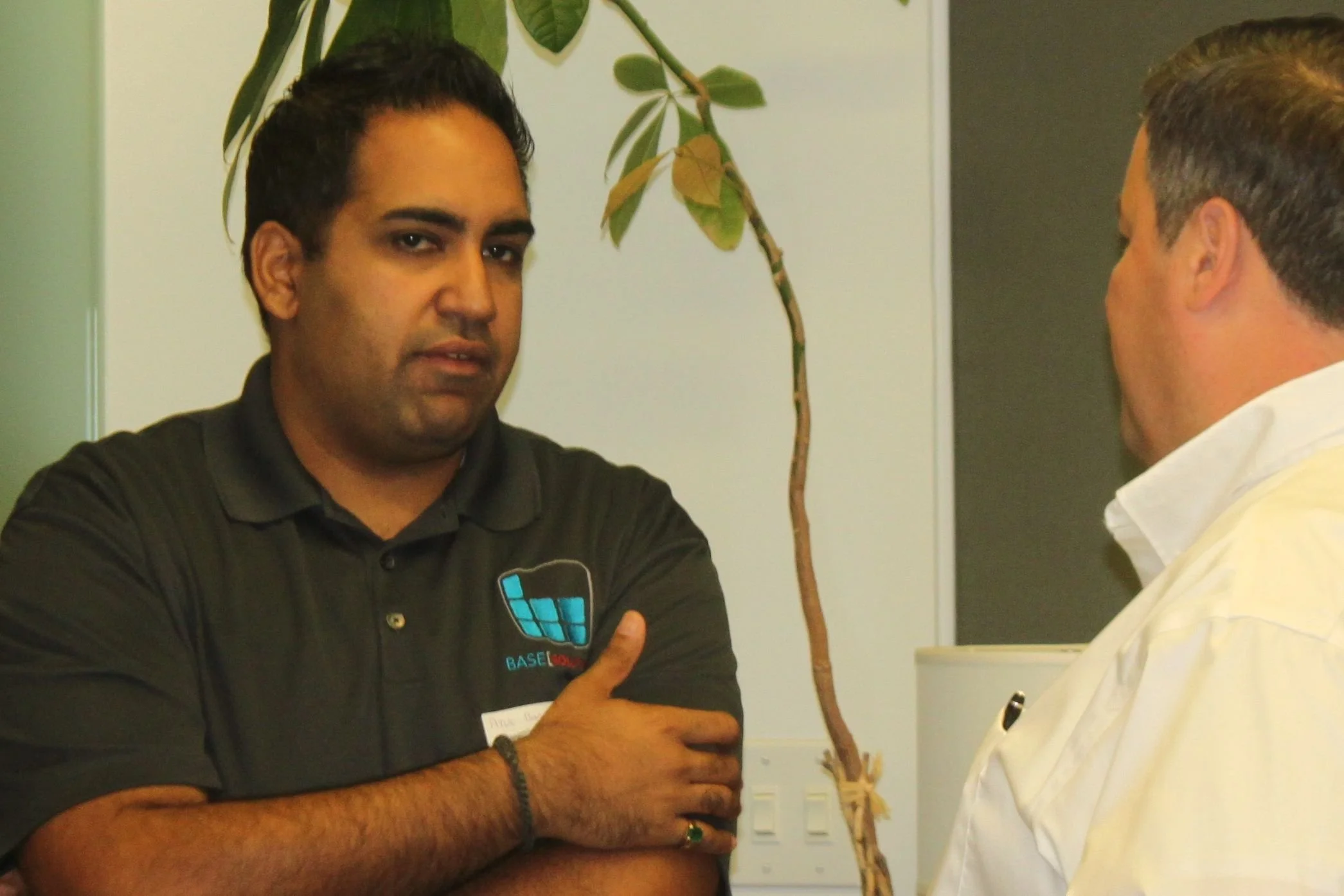 Two men are engaged in a conversation, one with crossed arms wearing a black polo shirt with a logo, and the other with a white shirt, in an indoor setting with a potted plant in the background.