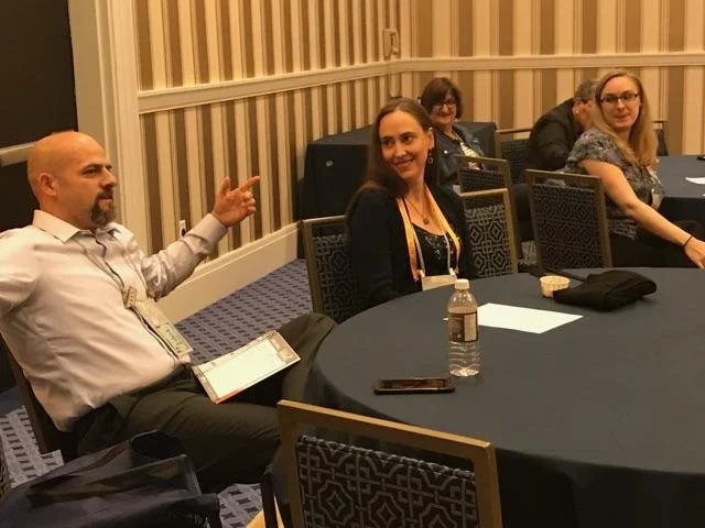 Four people sitting at a round table in a conference room, with one man gesturing while the others listen. The table has a water bottle, papers, and a phone.