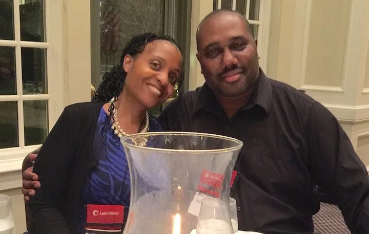 Two people sitting closely together at a table, smiling, with a large glass pitcher and glasses in front of them, indoors near windows with white framing.