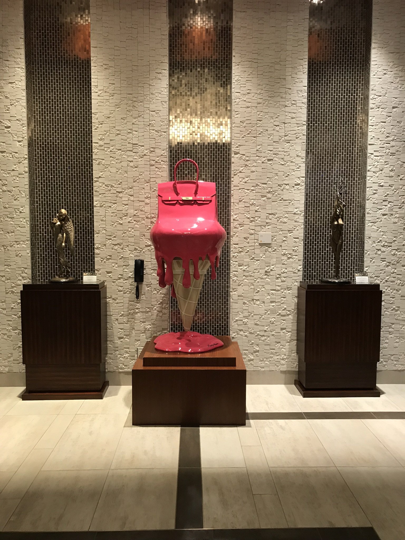 Decorative display with a bright pink handbag on top of an ice cream cone sculpture, flanked by two small statues on pedestals, with a textured wall background.